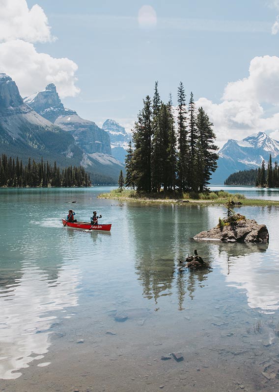 Two people canoe on a lake near a small tree-covered peninsula.