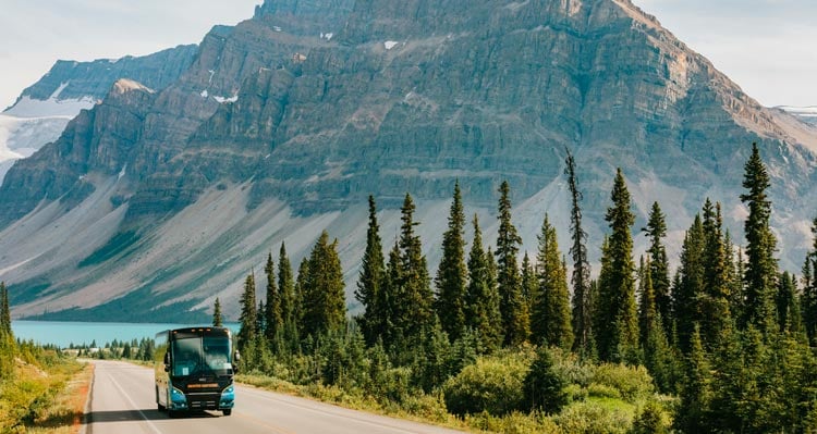 A bus drives along a forest-edged road below mountains.