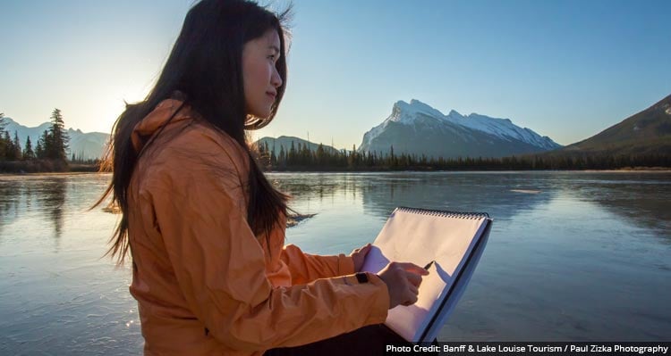 A woman sits near a lake with a drawing pad.