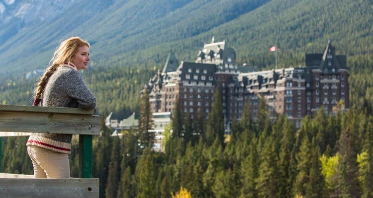 A woman stands at a wooden railing looking towards the Banff Springs Hotel