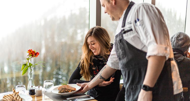 A chef brings a dinner to a window-side table.