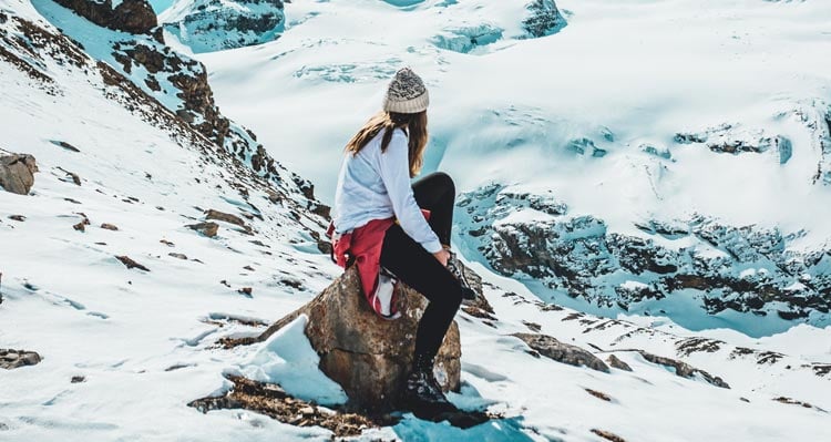 A person sits on a rock atop a snowy mountain landscape.