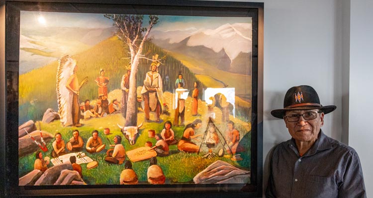 Roland Rollinmud stands in front of his painting of people gathered around a whitebark pine tree