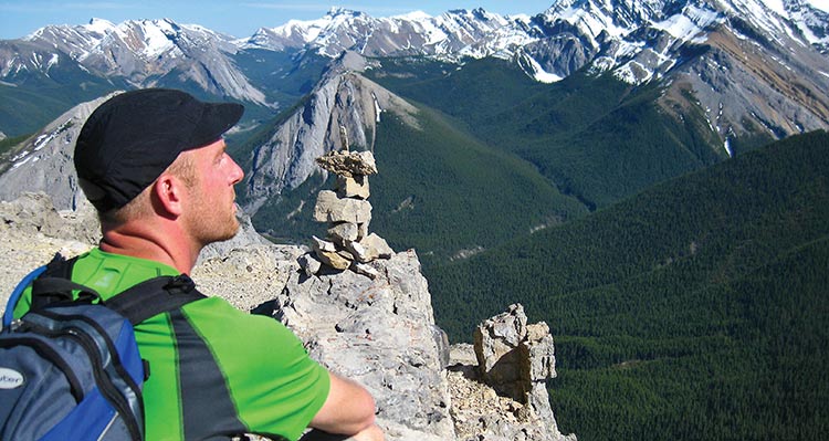 A hiker takes a rest atop a mountain, looking over wide forested valleys and rocky peaks.