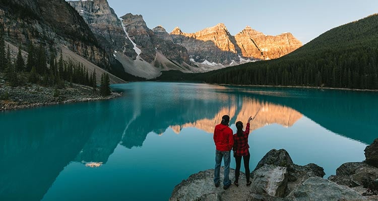 Two hikers stand on some rocks before a blue lake and jagged mountain peaks.