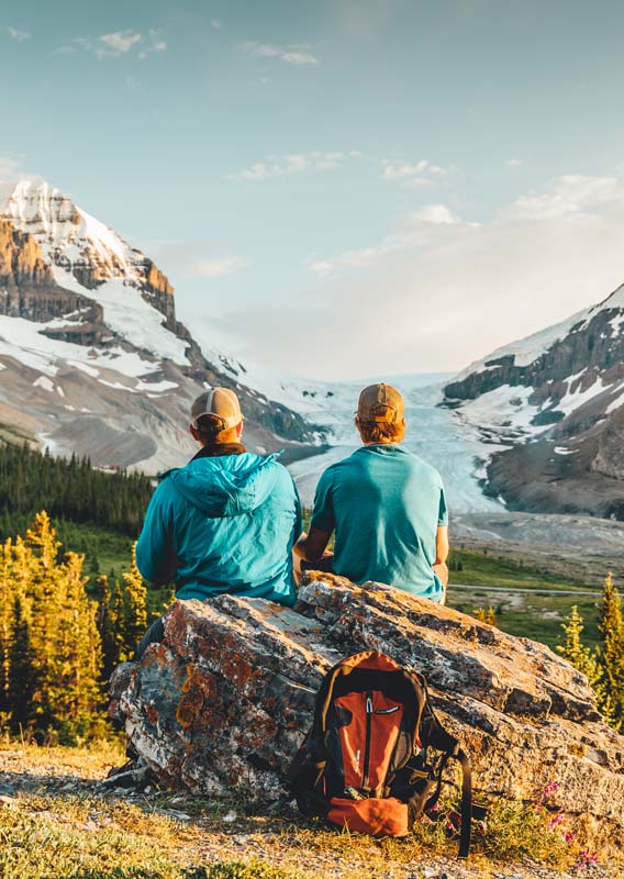 Two hikers take a break on a rock next to a forest, looking towards the Athabasca Glacier