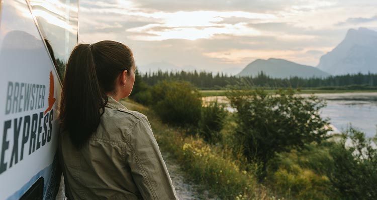 A woman leans against a bus looking towards a mountain and lake view.