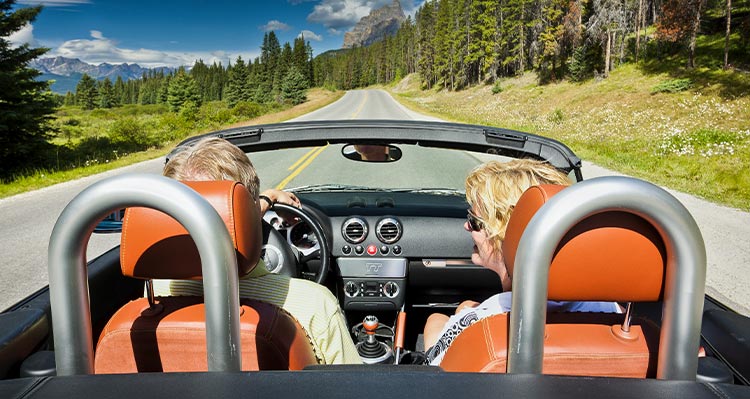 Two people drive in a car down an open road between conifer trees.