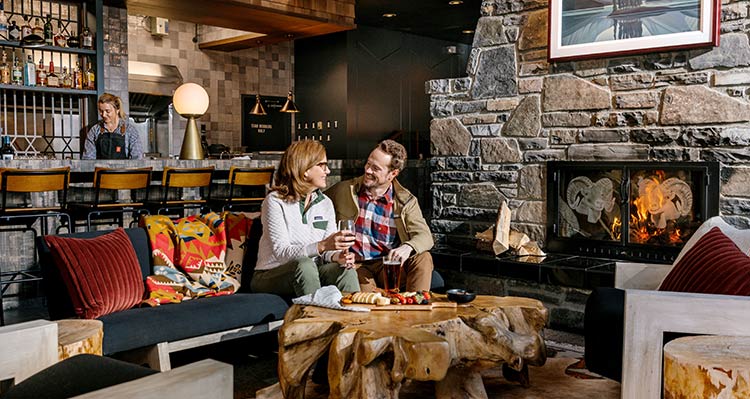 Two people sit at a table next to a large stone fireplace in a rustic restaurant