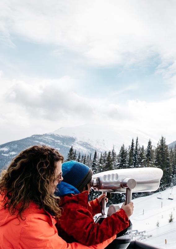 A mom and child look through sightseeing binoculars at a ski resort.