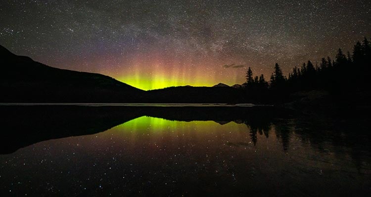 A night time view of a lake, with northern lights in the distance behind a mountain range.