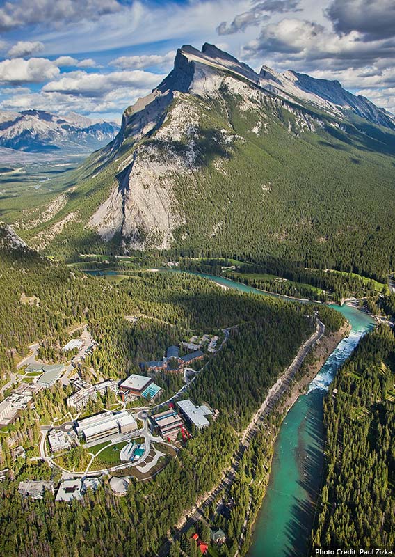 An aerial view of Banff, with Mt. Rundle rising behind a river winding through the forest.