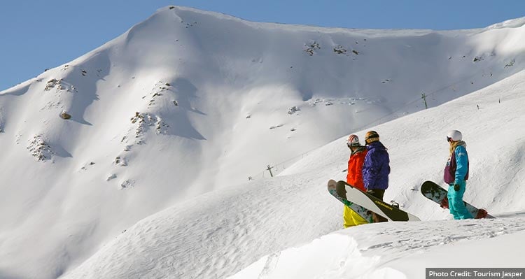 Three people with snowboards look down a run at a ski resort