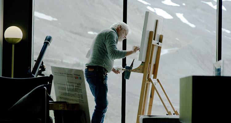 A painter stands at an easel, painting in front of a wide window looking out towards mountains.