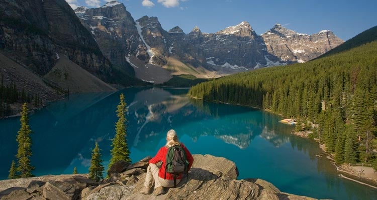 A person sits atop a rock pile overlooking a blue lake surrounded by high mountains.