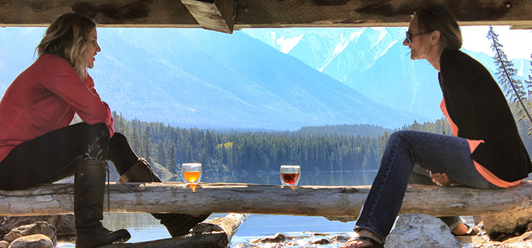 two women sit on a log by a lake and converse and enjoy tea