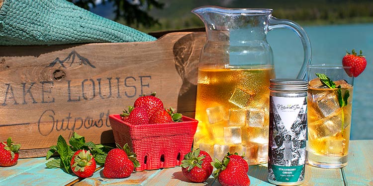 a pitcher with iced tea and strawberries near a lake and mountains in distance