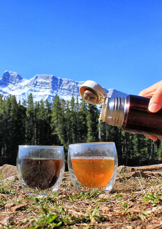 two clear tea mugs sit on the grass and a person pours tea into them