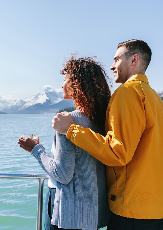 a couple stands at the edge of a boat deck drinking cocktails and looking out over water