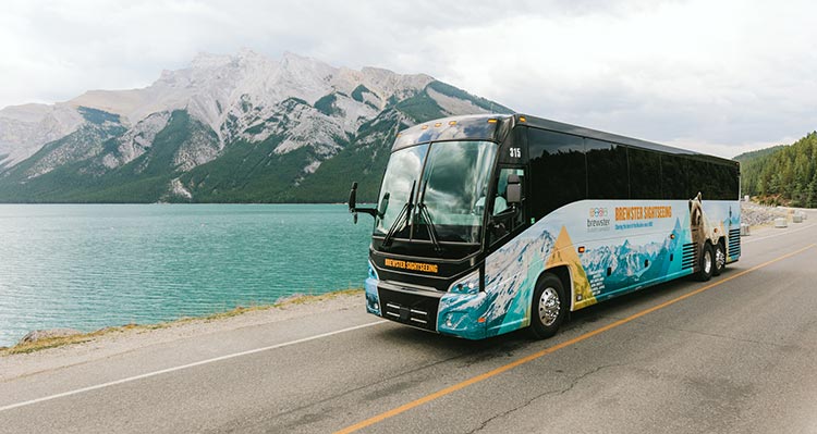 A bus drives along a road next to a large lake below a tall mountain.