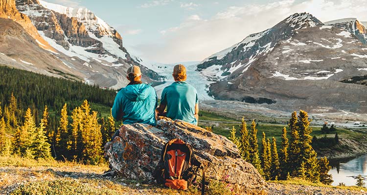 Two people sit on a rock, looking towards a glacier between tall mountains.