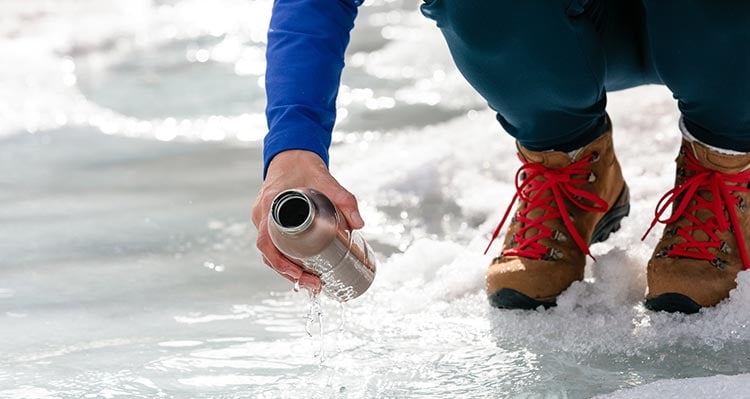 A person brings a water bottle down to water flowing from ice.