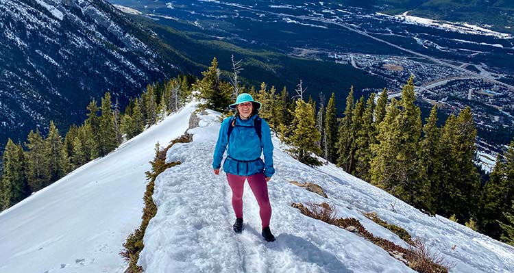 A hiker stands on a snowy path just above a stand a conifer trees.