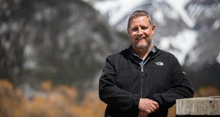 A bearded man leans against a picnic table.