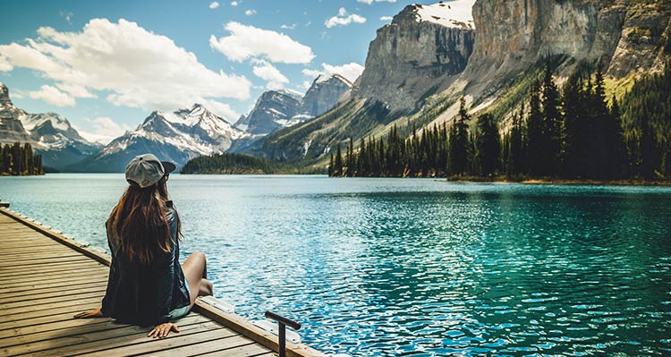 A person sits on a dock at the edge of the large blue lake below rising mountains.