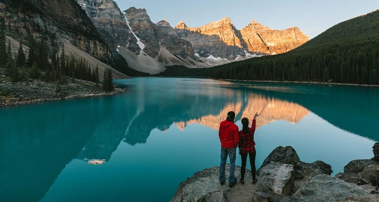 Two people look out across a blue lake below towering rocky peaks.
