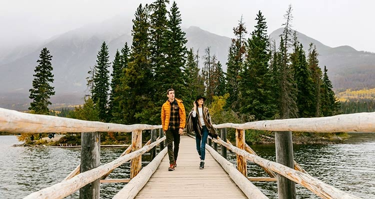 Two people walk on a wooden bridge from a small tree-covered island.