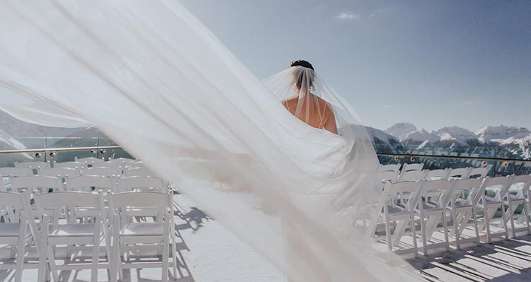 A bride walks down an aisle between chairs on a balcony overlooking mountains.