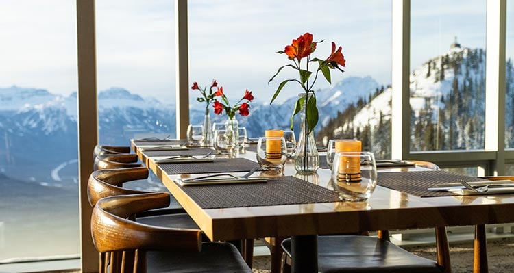 Dining tables set up with flowers along a tall window overlooking mountains and valleys.