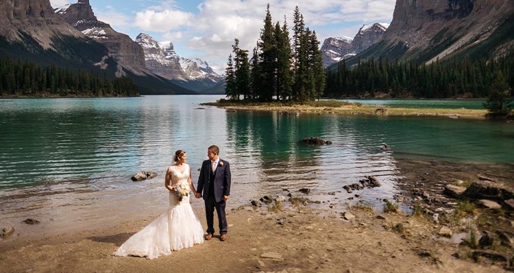 A bride and groom stand on the shore of a blue lake below tall mountains.