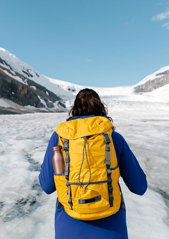 A person wearing a yellow backpack stands looking towards a glacier between mountains.