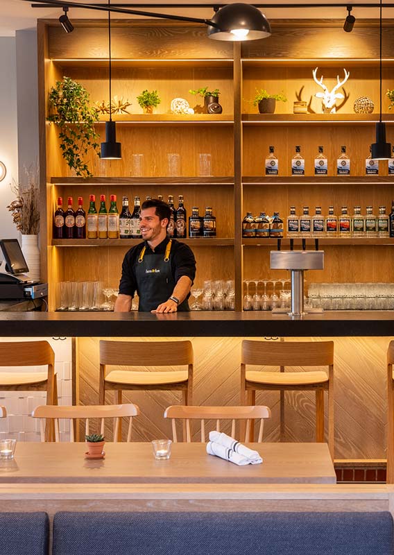 A bartender stands behind a bar decorated with light wood.