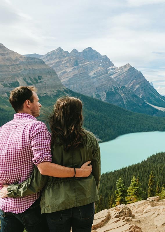 Two people stand overlooking a turquoise lake between forested mountains.