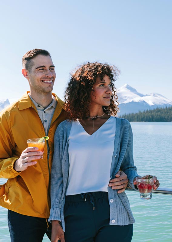 Two people stand on a boat looking on across a lake towards mountains.