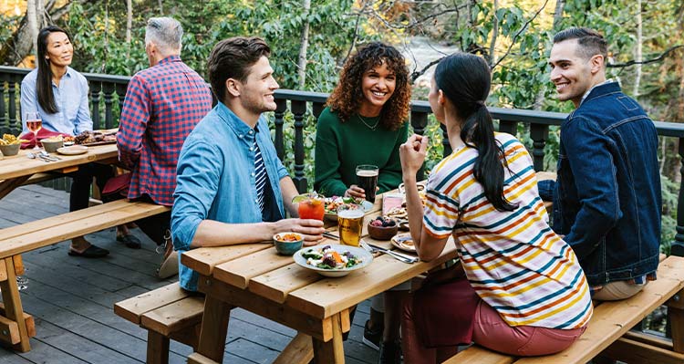 A group of friends eat on a patio next to a lush forest.