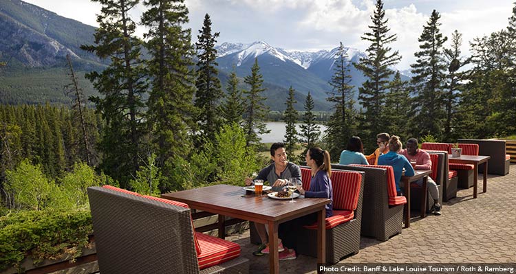 People sit at tables on a patio overlooking a forested valley between tree-covered mountains.