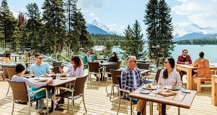 Groups of people sit at tables on a patio overlooking a large blue lake.