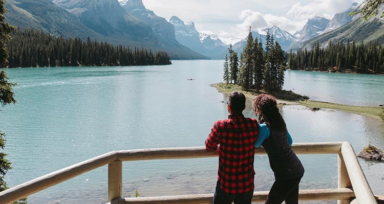 Two people stand at a viewpoint, looking out at a large mountain-sided lake.