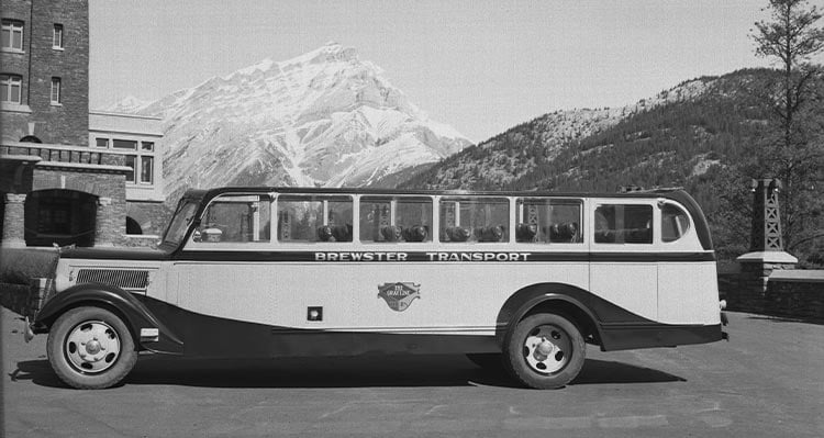 A historic photo of a tour bus parked in front of a stone building and snow-covered mountains.