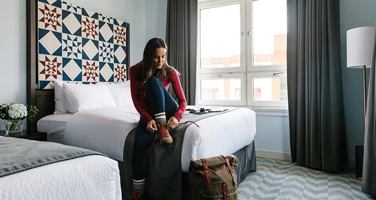 A woman puts on hiking boots, sitting at a bed's edge.