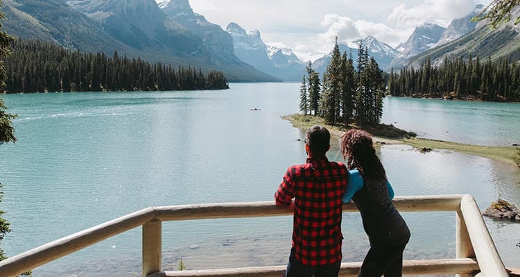 Two people stand at a lookout towards a lake below tall mountains.