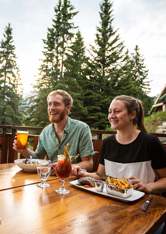 A group of people sit for dinner on a patio overlooking a conifer forest.