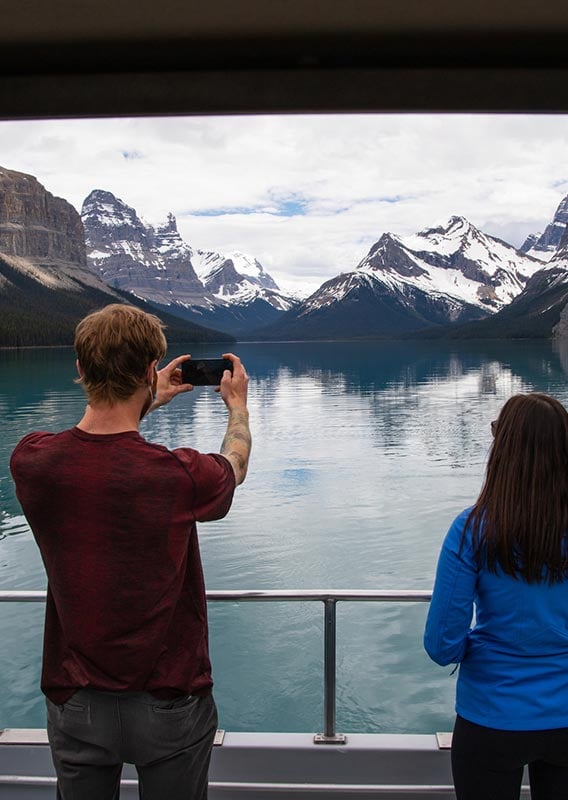 Two people stand at a boat's edge to take a photo of a mountain landscape.