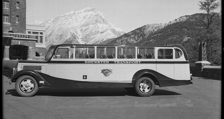 A historic photo of a tour bus parked in front of a stone building and snow-covered mountains.