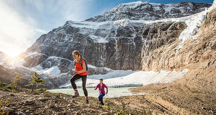 Two hikers walk on open terrain below snow-covered mountains.