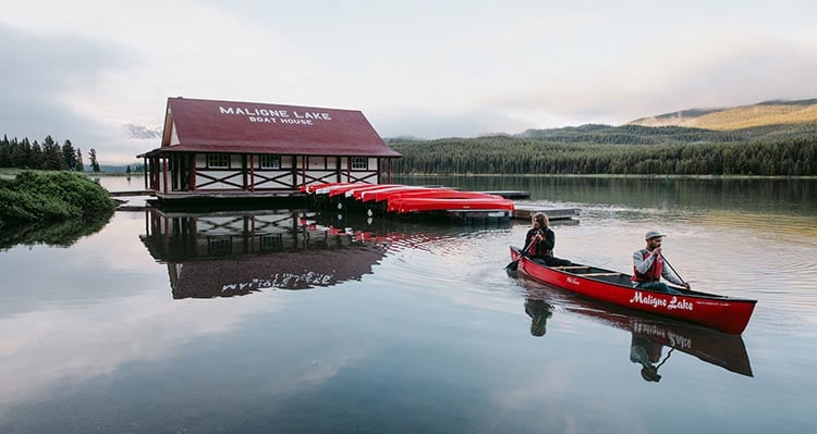 Two people canoe away from a lakeside boathouse.